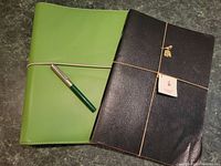 Two leather look binders/photo shows green and dark brown with elastic ties; green binder includes calligraphy pen attached, brown binder features artisan tag with book and bird design.