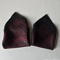 Two folded silk pocket scarves, one showing a paisley pattern and the other a small repeating diamond pattern, both in burgundy and black.