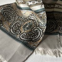 Close-up of scarf fabric showing intricate gold, taupe, green, and silvery paisley and ornamental designs with fringed edges.