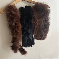 Three fur coat collars hanging on a wooden hanger against a background. Includes one reddish-brown fox fur collar and two shorter rabbit fur collars, one black and one brown.