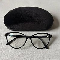 Black velvet Tom Ford eyeglasses case alongside the black-framed eyeglasses placed flat on a neutral fabric surface.