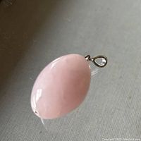 Close-up photo of the polished egg-shaped rose quartz pendant lying on a dark reflective surface, showing the stone and bail attachment.