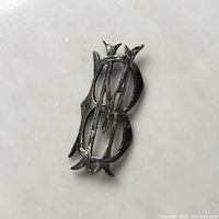 Front view of the sterling silver MB emblem pin showing ornate intertwined letters and patina visible on surface.