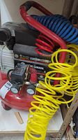 Side view of King Canada 2 HP air compressor showing red tank, gauges, regulator knob, and coiled yellow air hose.
