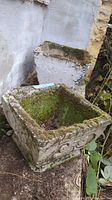 Two heavy concrete garden pots placed outdoors showing moss growth and wear. One pot is square with decorative scrolls, the other is rectangular with corner damage.
