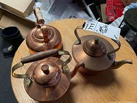 Photo showing three vintage copper tea pots placed on a table, highlighting the wooden grip handle tea pot and the brass-tone handle pot.