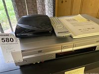 Front view of silver Sony combo DVD player and VHS VCR showing front AV inputs and controls with remote and telephone answering machine on top, instruction manual beside.