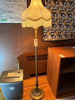 Full view of metal base floor lamp with cream fabric shade with fringe, standing on wooden floor against wall.