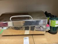 Side view of Kuuma stainless steel table top BBQ showing lid handle and propane tanks attached.