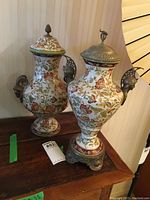 Two tall Wong Lee urns with floral porcelain bodies, ornate metal handles resembling animal heads, metal lids, and detailed metal bases on wooden surface.
