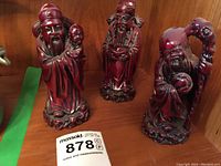 Front view of three red resin Chinese god figurines arranged on a wooden shelf.
