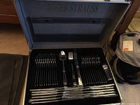 Open aluminum case with black foam insert showing two stacks of dinner forks on left, two stacks of teaspoons on right, three display utensils in center and multiple wrapped knives in bottom row