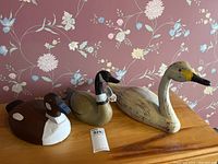 Three assorted wooden decoy ducks on a wooden surface against floral wallpaper background. Shows size comparison and general wear.