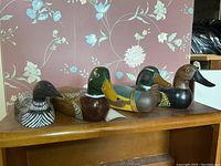 Four assorted wooden duck decoys displayed on a wooden surface against floral wallpaper. Varied paint colors and wear visible.