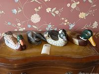 Four assorted wooden decoy ducks arrayed on wooden surface against floral wallpaper background, showing overall set and size range.