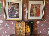 Two framed Klimt prints hanging on floral wallpaper with Klimt 6 posters box below