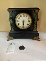 Front view of black wood mantle clock with decorative gold-tone metal feet, faux marble columns, and glass face showing brand on dial.