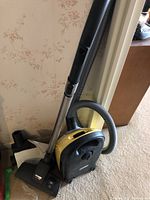 Photo showing the yellow and black Miele vacuum cleaner standing upright with hose and metal wand attached.
