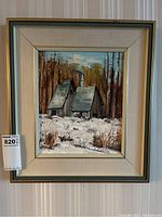 Framed original painting of a winter rural scene with buildings and trees, mounted on a striped wall.