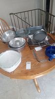 Photo of set of glass and metal bakeware on wooden table showing multiple baking and roasting pans, including clear and blue glass dishes, metal broiler pan with rack, round aluminum pans and tube cakes pans.