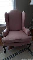 Front view of an upholstered wingback chair in light pink fabric with arm covers and dark wood cabriole legs.