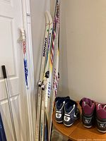 Cross country skis including Salomon Atomic, Bonna, and Atomic Touring leaning against a wall with ski boots next to them.