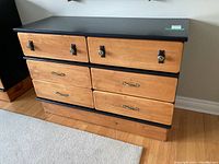 Front view of wooden 6-drawer dresser showing black painted top, frame and natural wood drawer fronts with mixed metal hardware handles.