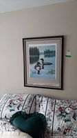 Full view of framed wall art hanging above a floral patterned couch, showing ducks on a lake with trees in the background.