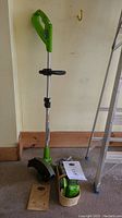Full view of Greenworks cordless string trimmer standing upright next to a wall, with batteries, charger, and manual on the floor beside it.