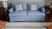 Front view of light blue Ethan Allen two-seater sofa with two plaid toss cushions on a rug between two side tables.