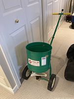 Full view of the green Spin-On broadcast spreader with wooden handle against a white door background.