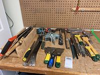 Assorted hand tools on workbench with hammers, scrapers, crowbars, clamps and drill bits, showing overall condition and variety.