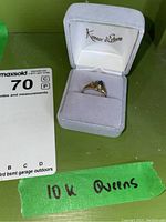 Ring displayed in gray velvet box with Kimaar d'Estee branding. Green tape labeled '10k Queens' visible.