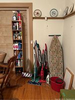 Full view of corner with vacuum cleaners, ironing board, red mop bucket, iron, and wooden cabinet filled with cleaning supplies.