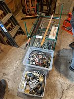 Photo of metal rack, carpenter's squares, large chain with hook, wooden ruler and two plastic bins filled with assorted cords and tools on floor in garage.