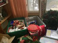 Overview of boxes and bins containing multiple Christmas decorations including pot holders, pinecone wreaths, and a red pillow with 'PEACE'.