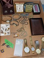 Overview of all items in lot showing clocks, watches, wallet, necklaces, cuff links, tie clips, and pins arranged in a cardboard box.