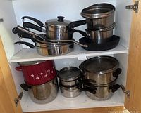 View of upper and lower shelves in cabinet showing various pots, pans and saucepans stacked neatly, including stainless steel and one red enamel pot with floral design.
