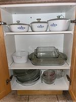 Full cabinet view with Corning Ware on top, glass bakeware and pie plates in the middle and bottom shelves, plus two glass measuring cups on bottom shelf.