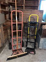 Front view showing the push broom leaning against the red 4-wheel dolly and black and yellow 2-wheel dolly in shed environment.