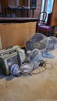 Wide angle shot showing all fans and heaters in lot with furniture in background. Items include oscillating table fans, clip-on fans, and two vintage Torcan heaters.