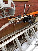 Photo showing an electric tree saw with metal frame and motor alongside two Fiskars manual tree saws with orange handles and curved blades, leaning against a brick wall above a ladder with snow on ground.