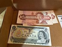 First photo showing two Canadian two-dollar bills and one Canadian one-dollar bill in a cardboard box.