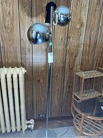Full view of tall chrome floor lamp with two spherical shades against wood panel wall, showing the entire lamp and base.