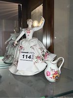 Porcelain figurine and floral pitcher are displayed side by side in a glass cabinet, showing the elaborate floral patterns and fine porcelain detail.