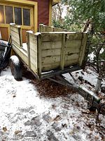 Overall view of the wooden utility trailer showing the weathered wood panels, black tires, and metal frame under snow and leaves.