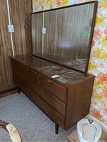 Front and angled view of vintage wooden dresser with 7 drawers and attached rectangular wood-framed mirror, showing surface wear and vintage wood paneling backdrop.