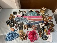Full lot of costume jewelry laid out on white surface showing variety of necklaces, earrings, bracelets, and brooches in assorted colors.