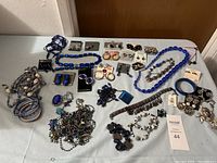 Full view of assorted costume jewelry laid out on table, mainly blue and metallic tones, featuring necklaces, earrings, bracelets, rings, and brooches.