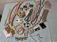 Photo showing assorted pearl and bead necklaces, bracelets, earrings, gold-tone chain necklace, and various costume jewelry pieces on a gray textile surface.